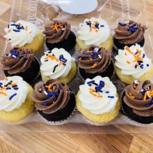 Assortment of a dozen mini cupcakes, chocolate cake with chocolate frosting and vanilla cake with vanilla frosting, with orange, white, black, and purple jimmies on top.