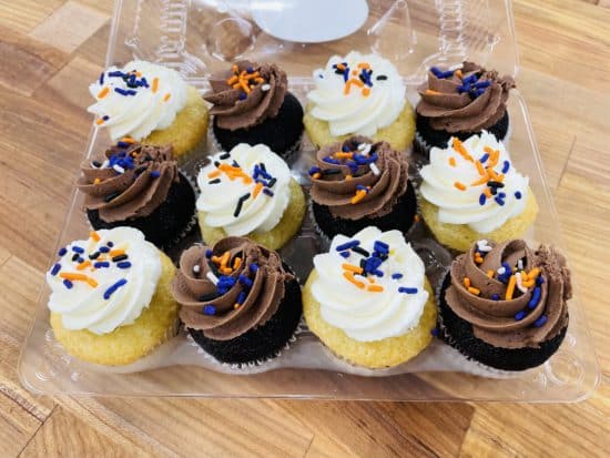 Assortment of a dozen mini cupcakes, chocolate cake with chocolate frosting and vanilla cake with vanilla frosting, with orange, white, black, and purple jimmies on top.