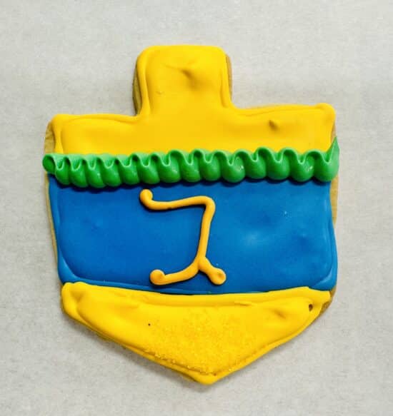 Royal iced dreidel cookie. Yellow top and bottom, a rippled green band at the top, and a blue band in the middle. In yellow frosting is the Hebrew letter "gimel".