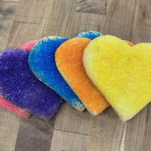 An array of large sanding sugar heart cookies showing multiple colors on a wooden countertop. Pink, purple, light blue, orange, and yellow are shown.