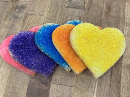 An array of large sanding sugar heart cookies showing multiple colors on a wooden countertop. Pink, purple, light blue, orange, and yellow are shown.