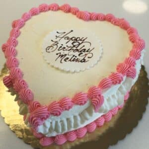 White heart-shaped cake with pink border decoration, on a gold cake board. A white oval plaque of fondant says "Happy Birthday Melissa" in chocolate script.